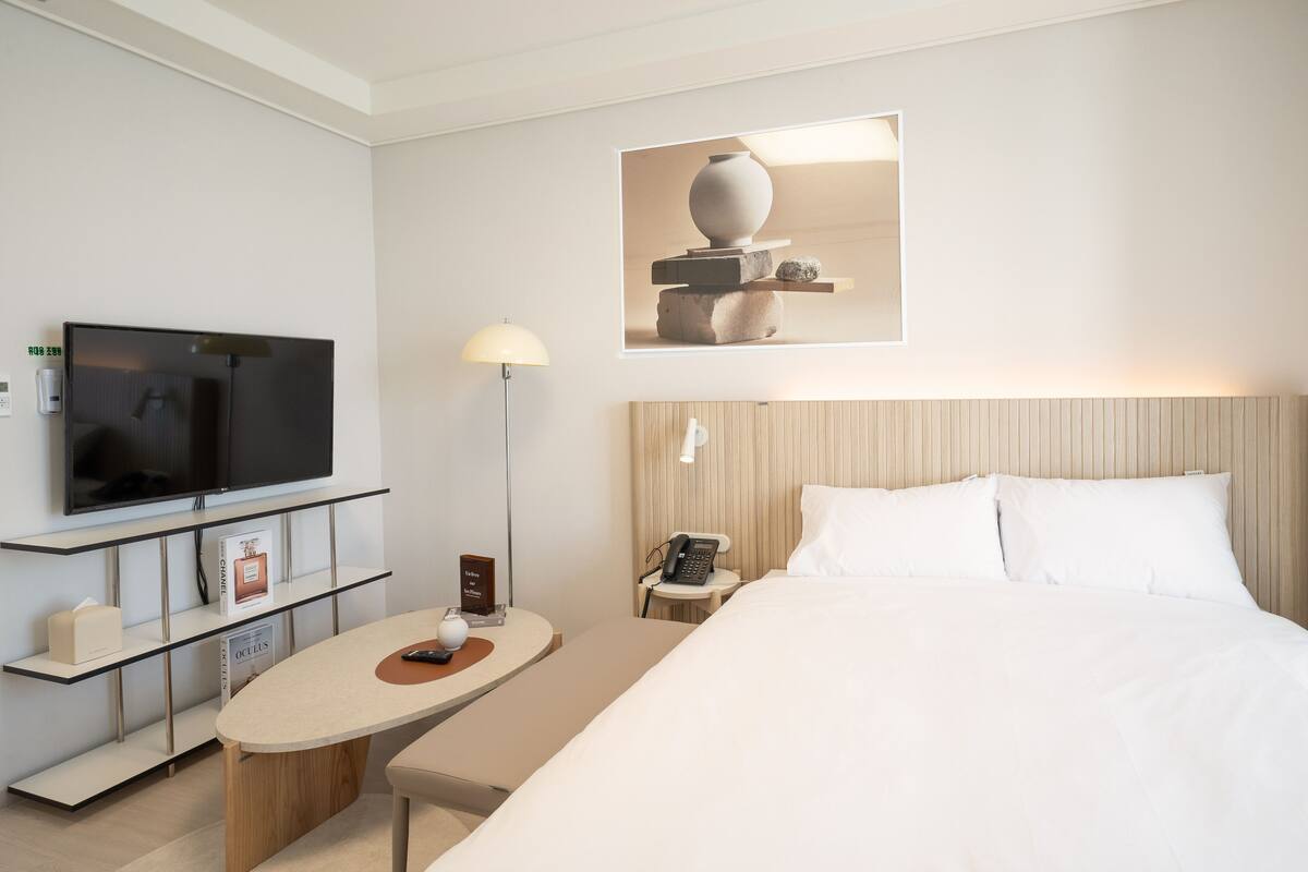 A modern hotel room with a large bed, a flat-screen TV, a bedside table with a lamp, a small desk with a chair, and a shelf with books and a vase.