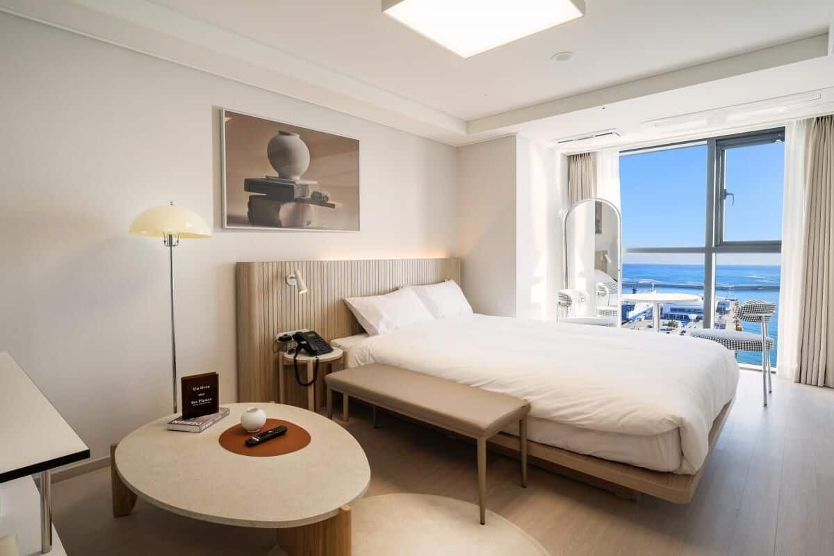 A hotel room with a large bed, a bedside table with a lamp, a small round table, a bench, a view of the sea, and a painting on the wall.