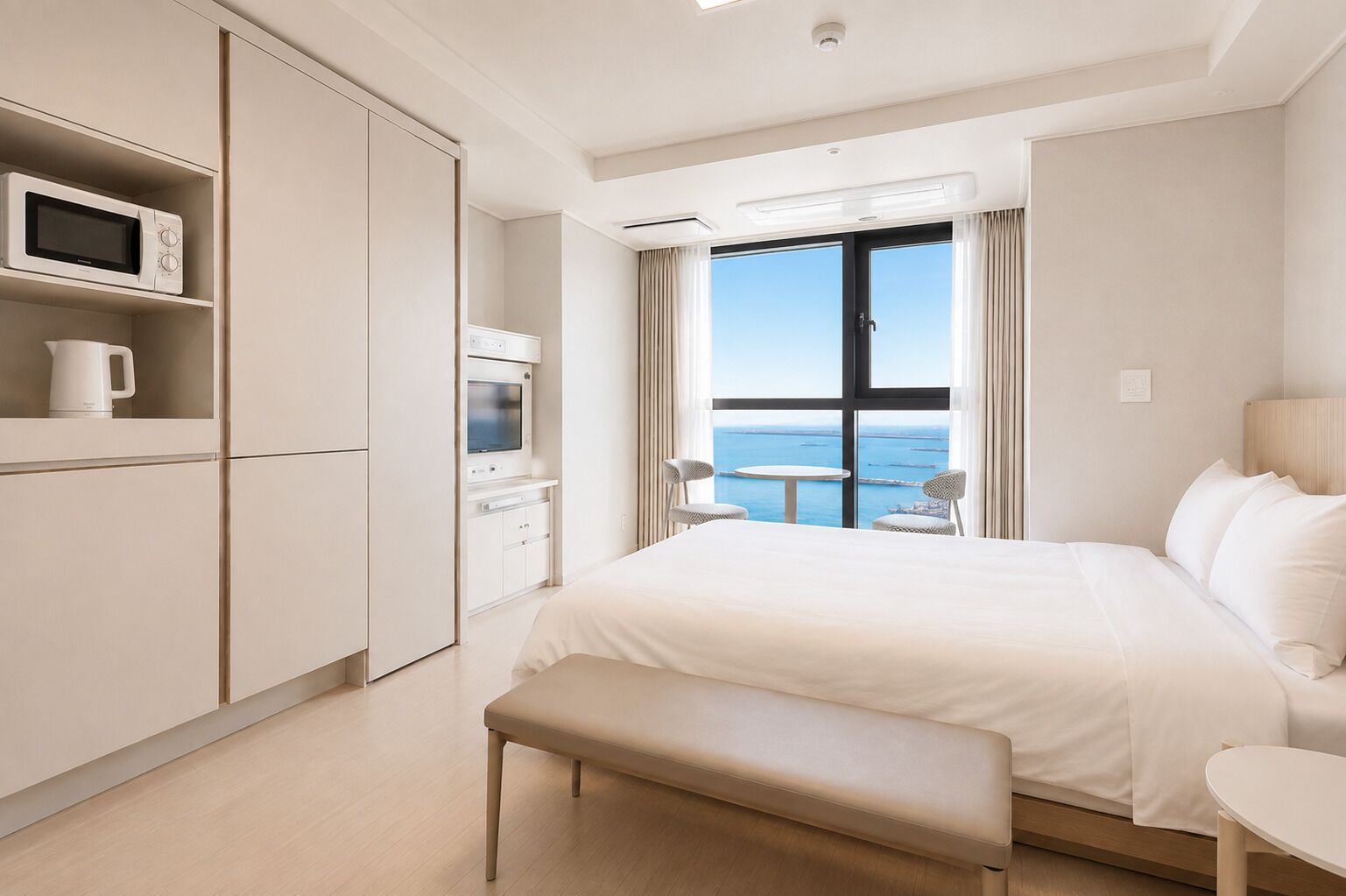 A hotel room with a bed, a bench, a TV, a microwave, and a view of the sea.