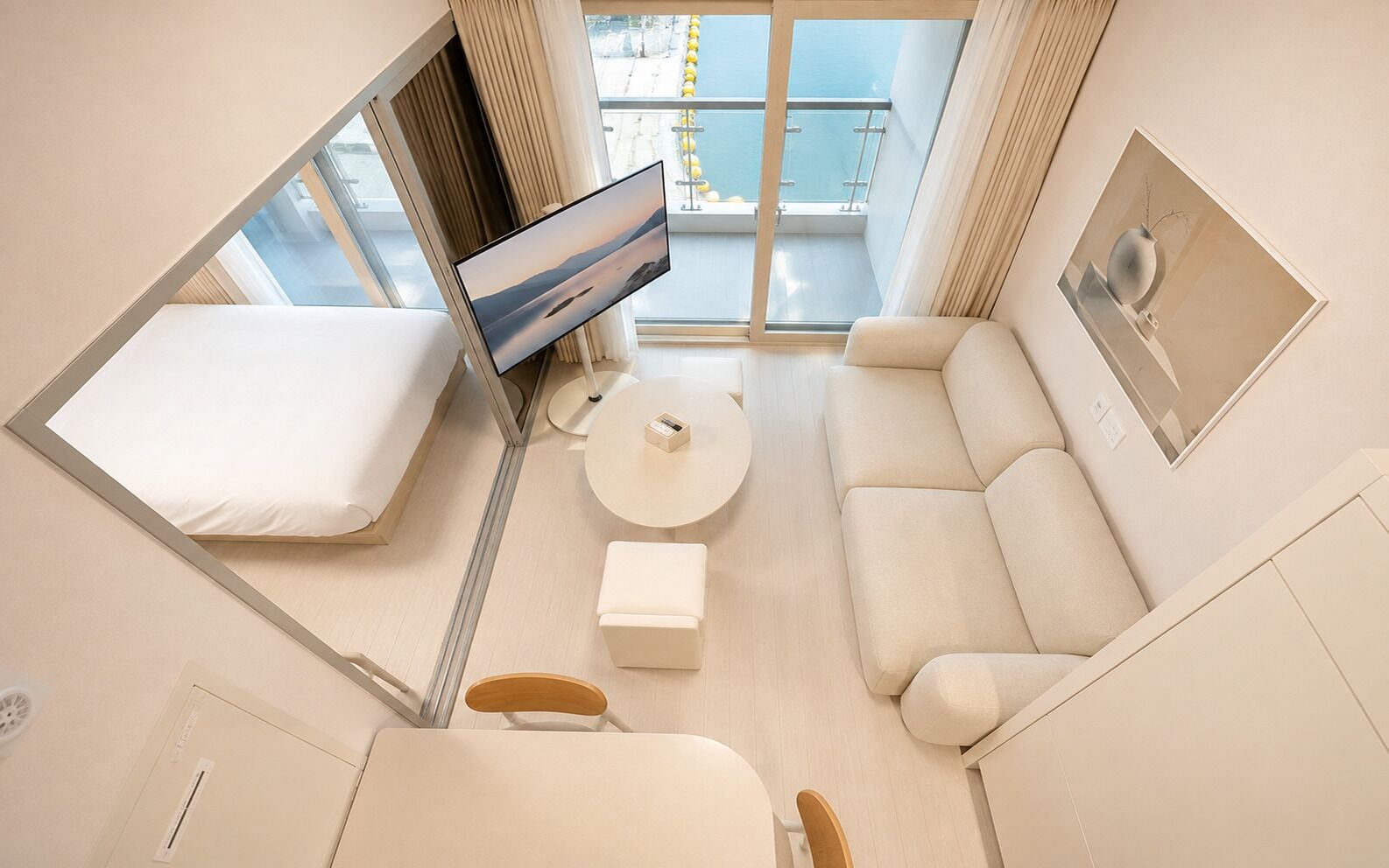 A modern hotel room with a bed, a sofa, a small table, and a TV. There is a balcony with a view of the sea.