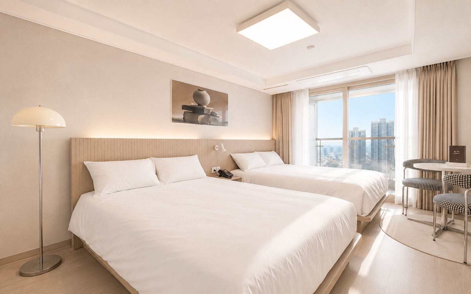 A hotel room with two beds, a desk with chairs, a lamp, and a city view.