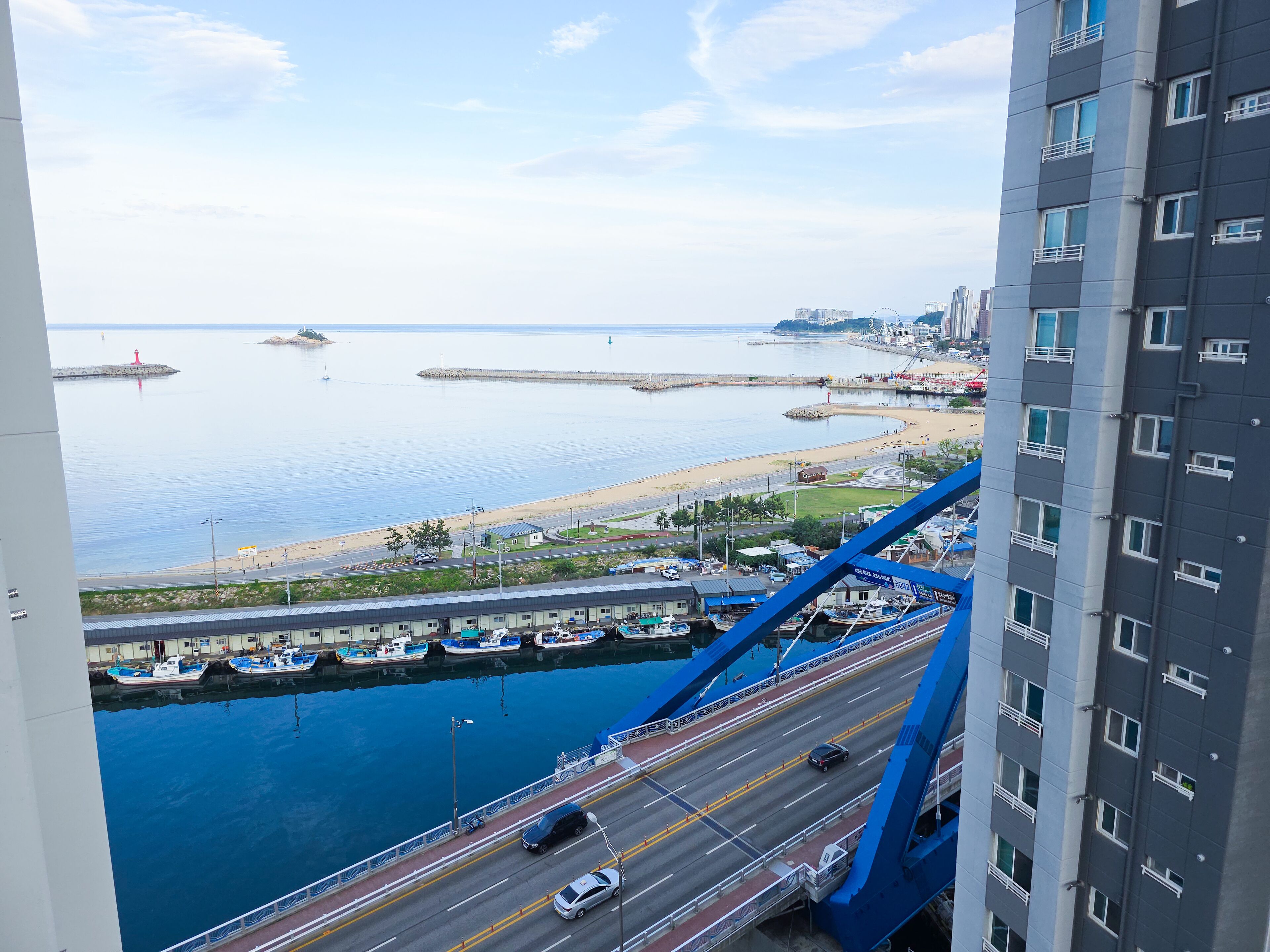 A high-rise building overlooking a marina with boats docked, a road with vehicles, and a beach in the background.