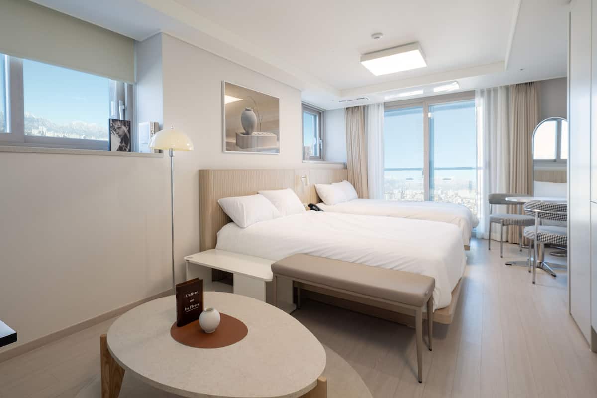 A hotel room with two beds, a round table with a vase, a bedside lamp, and a view of the city.
