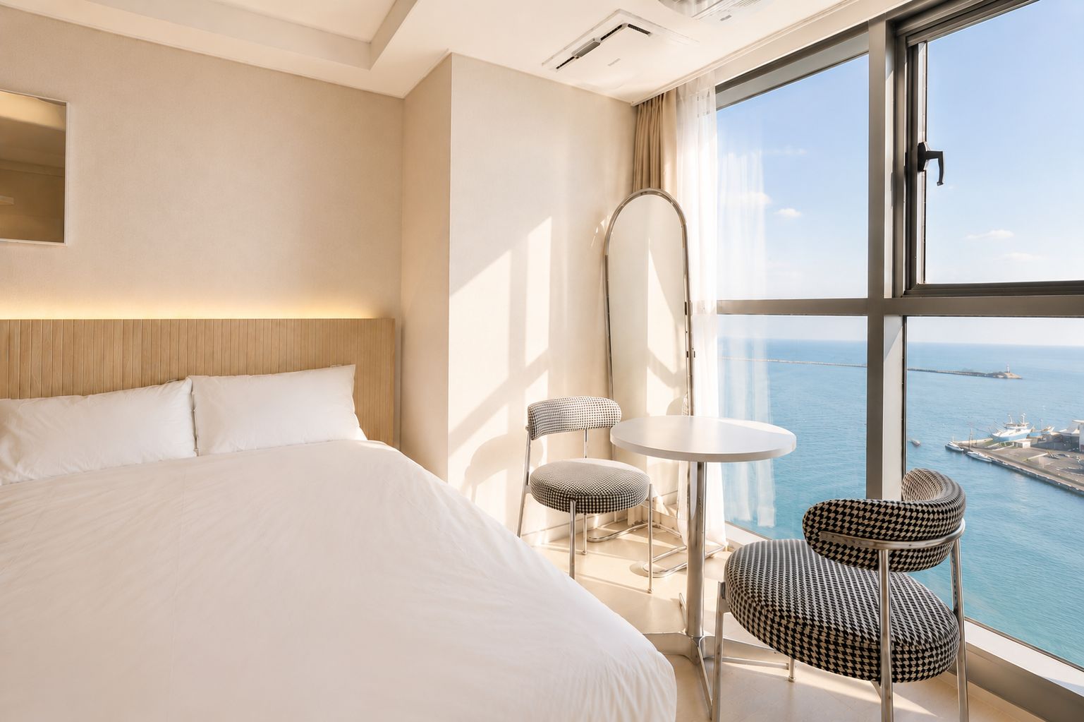 A hotel room with a large bed, a round table, two chairs, a mirror, and a view of the sea.