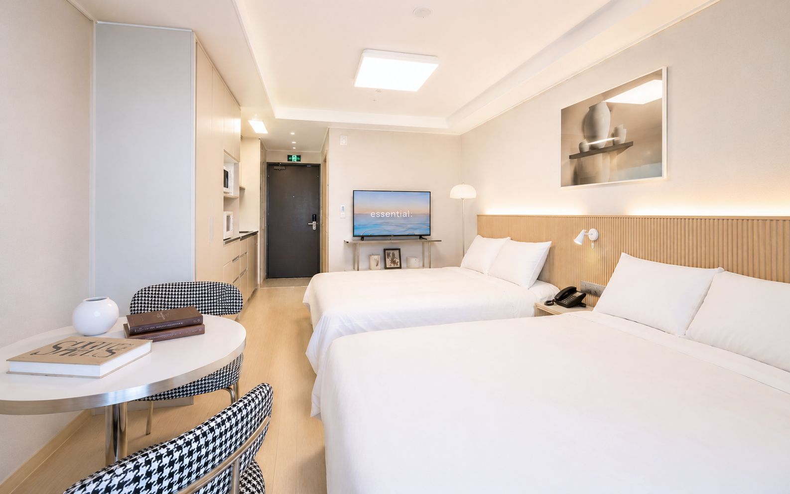 A hotel room with two beds, a small table with a chair, a TV, and a bedside table with a lamp.