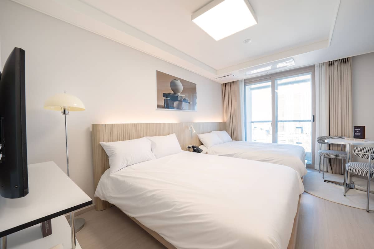 A hotel room with two beds, a desk, a chair, a lamp, and a window with curtains.