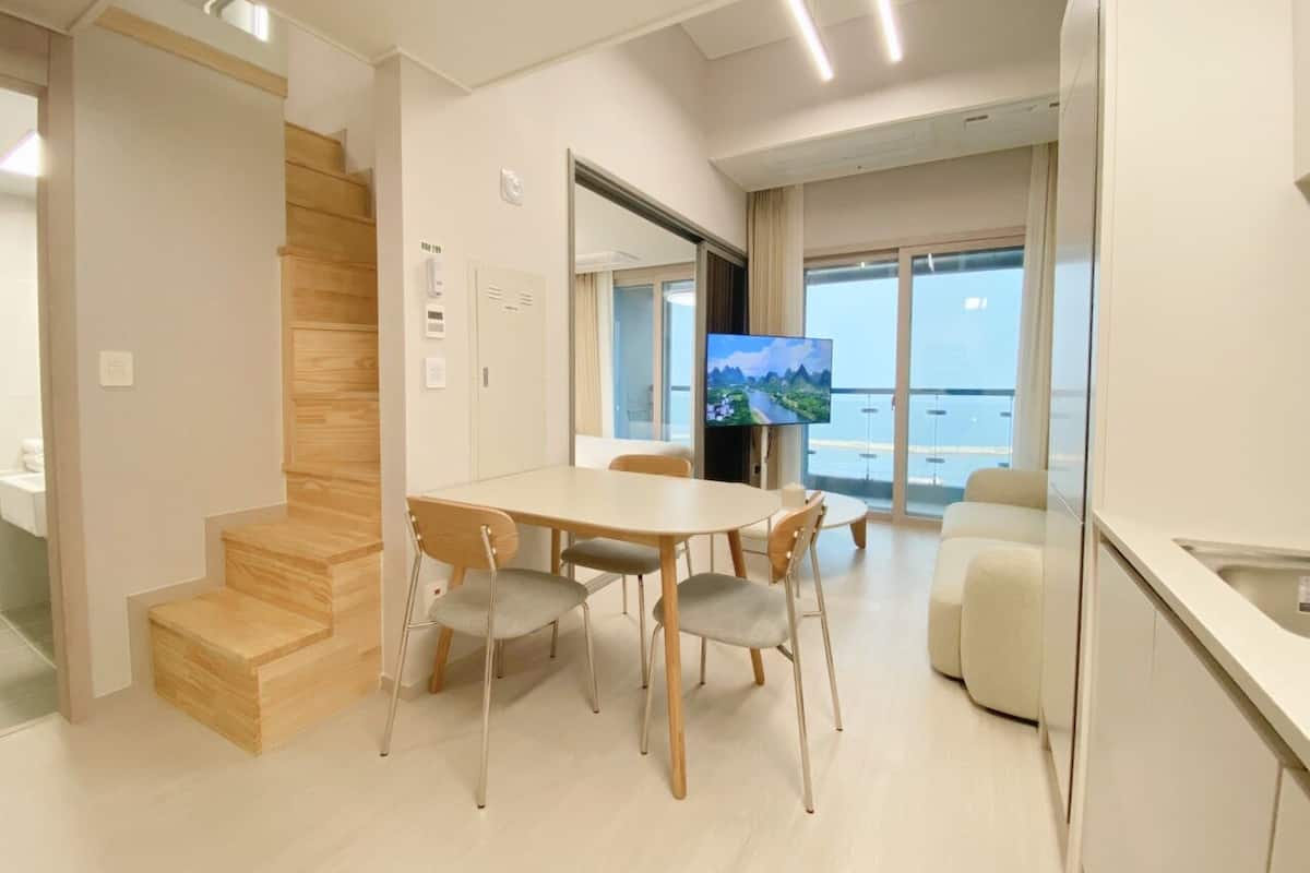 A modern kitchen with a dining area, wooden staircase, and a large window with a view.
