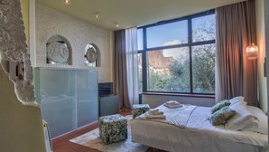 A bedroom with a large window, a bed with white bedding, a small table, and a decorative wall with a brick pattern.