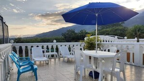 A terrace with white plastic chairs and tables, a blue umbrella, and a view of mountains. - Finca-Hostal Sanfelipe (Calima)