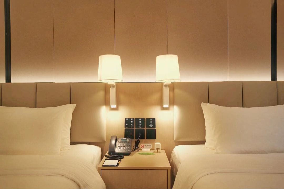 Habitación de hotel con dos camas, una mesita de noche con un teléfono, una lámpara y un cartel con instrucciones.