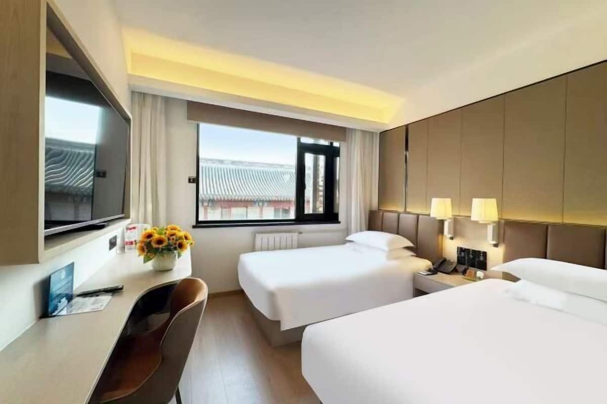 Habitación de hotel con dos camas, un escritorio con silla, un televisor y una ventana con vista a un edificio.