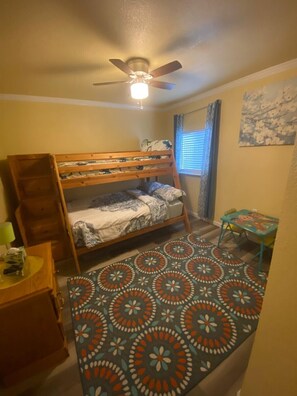 A small, cozy room with a bunk bed, a ceiling fan, a patterned rug, a small table, and a window with curtains.