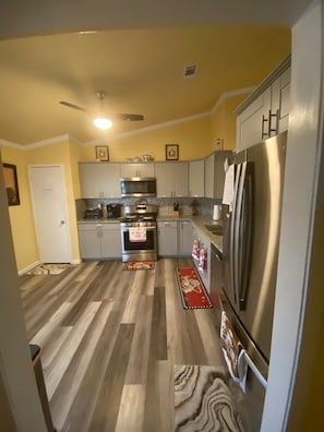 A kitchen with a stainless steel refrigerator, a microwave, and an oven. There are wooden floors and a ceiling fan.