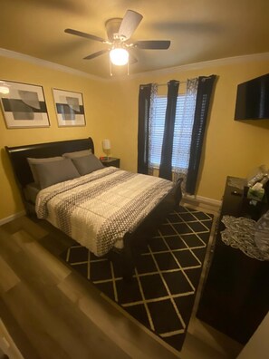 A bedroom with a bed, a ceiling fan, a nightstand, a dresser, and a window with curtains.