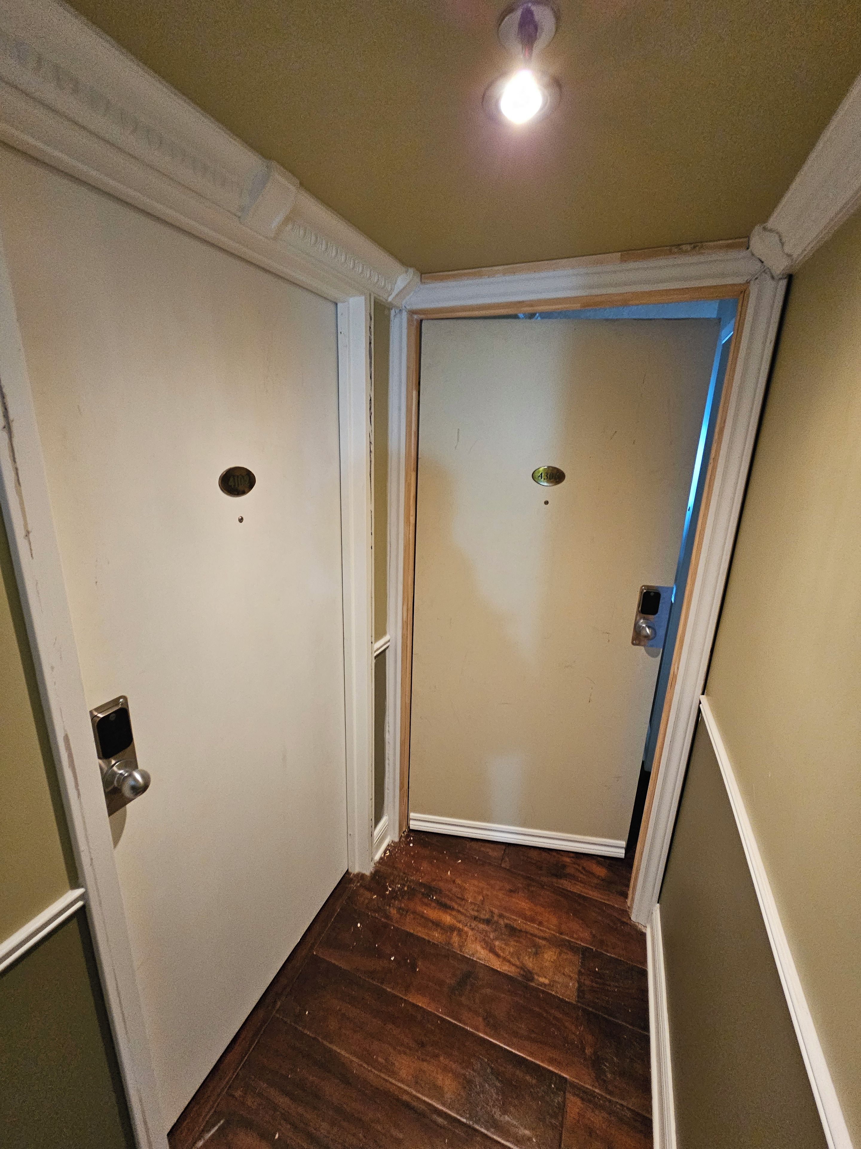 A narrow hallway with wooden flooring, white walls, and a door with a number plate.
