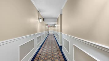 A long, narrow hallway with patterned carpet, white wainscoting, and wall-mounted lighting.