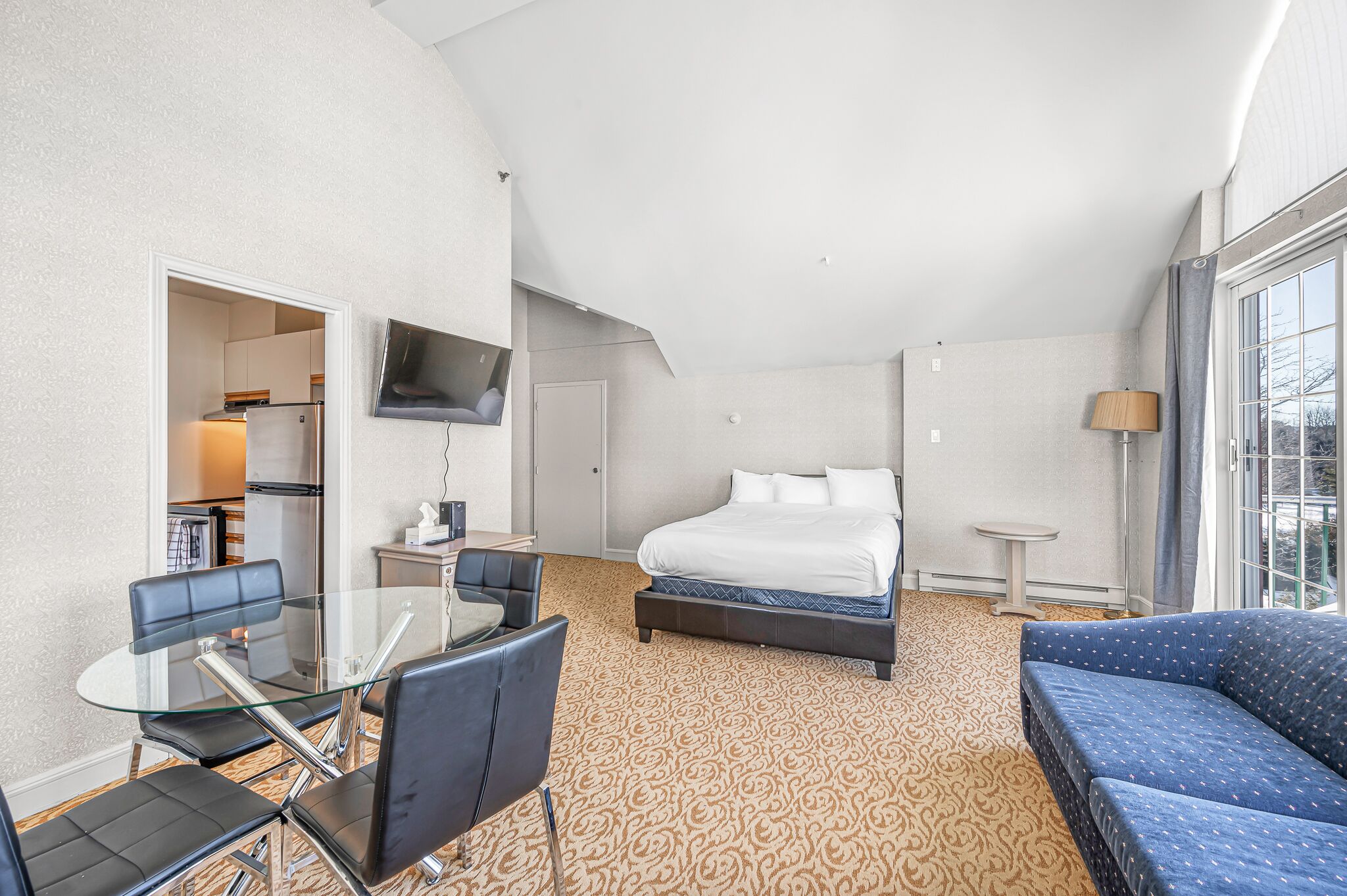 A hotel room with a bed, a sofa, a dining table with chairs, a TV mounted on the wall, and a window with curtains.