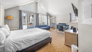 Habitación de hotel con una cama grande, un escritorio, una silla y un sofá. Tiene una ventana con cortinas, una lámpara y un televisor en la pared.