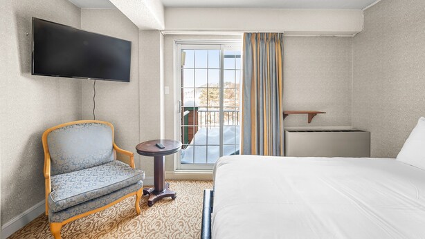 A hotel room with a bed, a chair, a TV mounted on the wall, a small table, a window with curtains, and a view of the outdoors.
