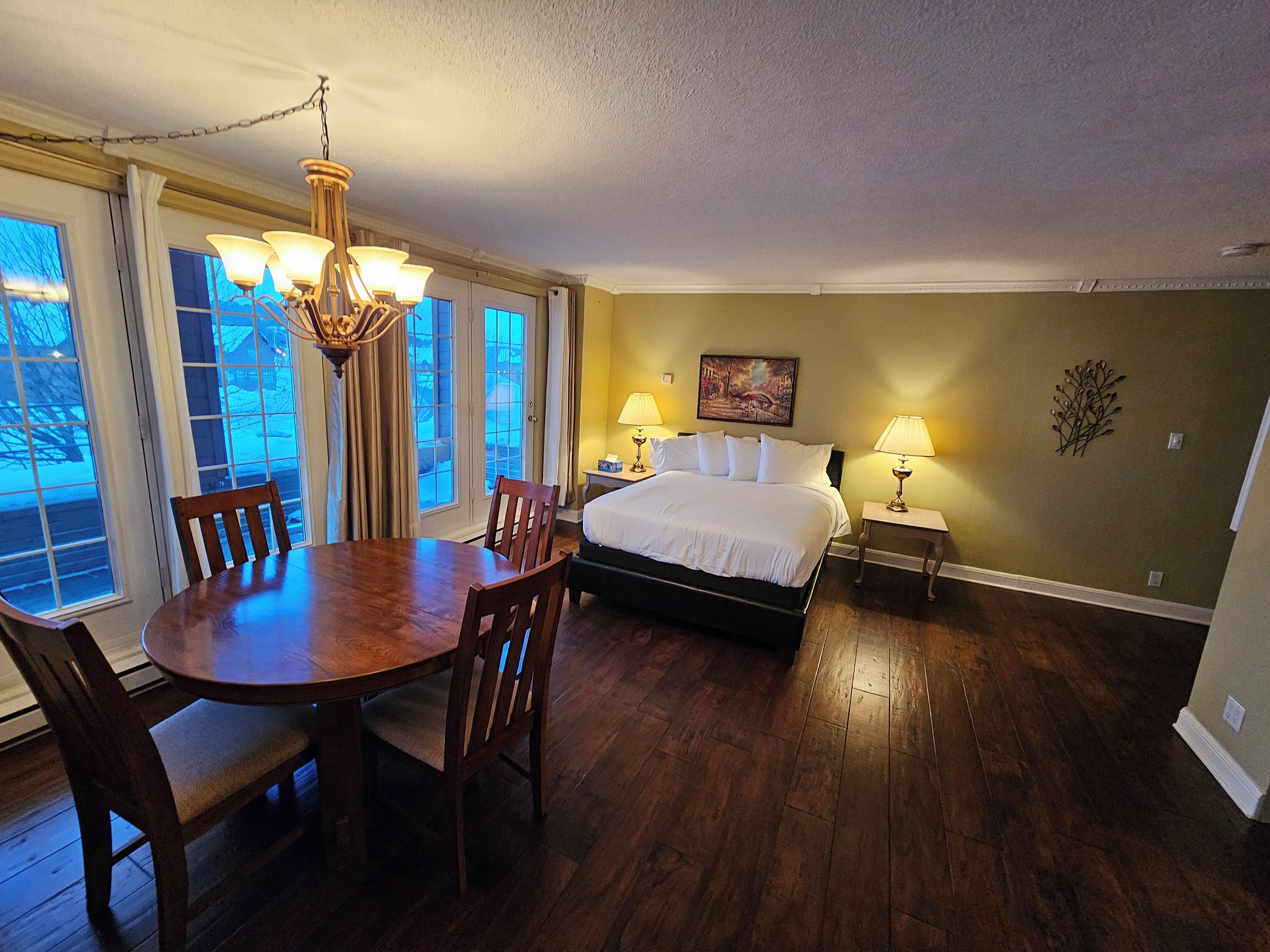 A room with a bed, a dining table with chairs, a chandelier, and a painting on the wall.