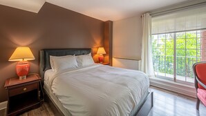 Une chambre d’hôtel avec un grand lit, deux lampes de chevet, une fenêtre avec des stores et une vue sur la verdure.