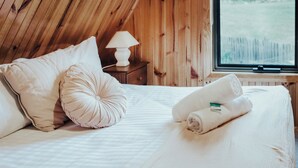 Una camera da letto con soffitto in legno, un letto con biancheria bianca, due cuscini, una lampada su un comodino e una finestra con vista sull'esterno.