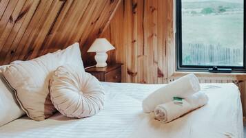 Une chambre avec un plafond en bois, un lit avec une literie blanche, deux oreillers, une lampe sur une table de chevet et une fenêtre donnant sur l’extérieur.