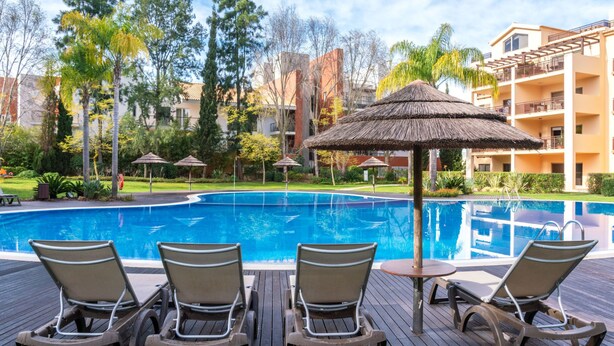 A swimming pool with lounge chairs and a thatched umbrella, surrounded by greenery and a multi-story building. - Triplex Vilamoura VIP Properties by Interhome (Vilamoura)