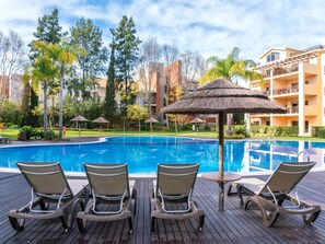 Una piscina con sillas de descanso y una sombrilla de paja, rodeada de vegetación y un edificio de varios pisos.