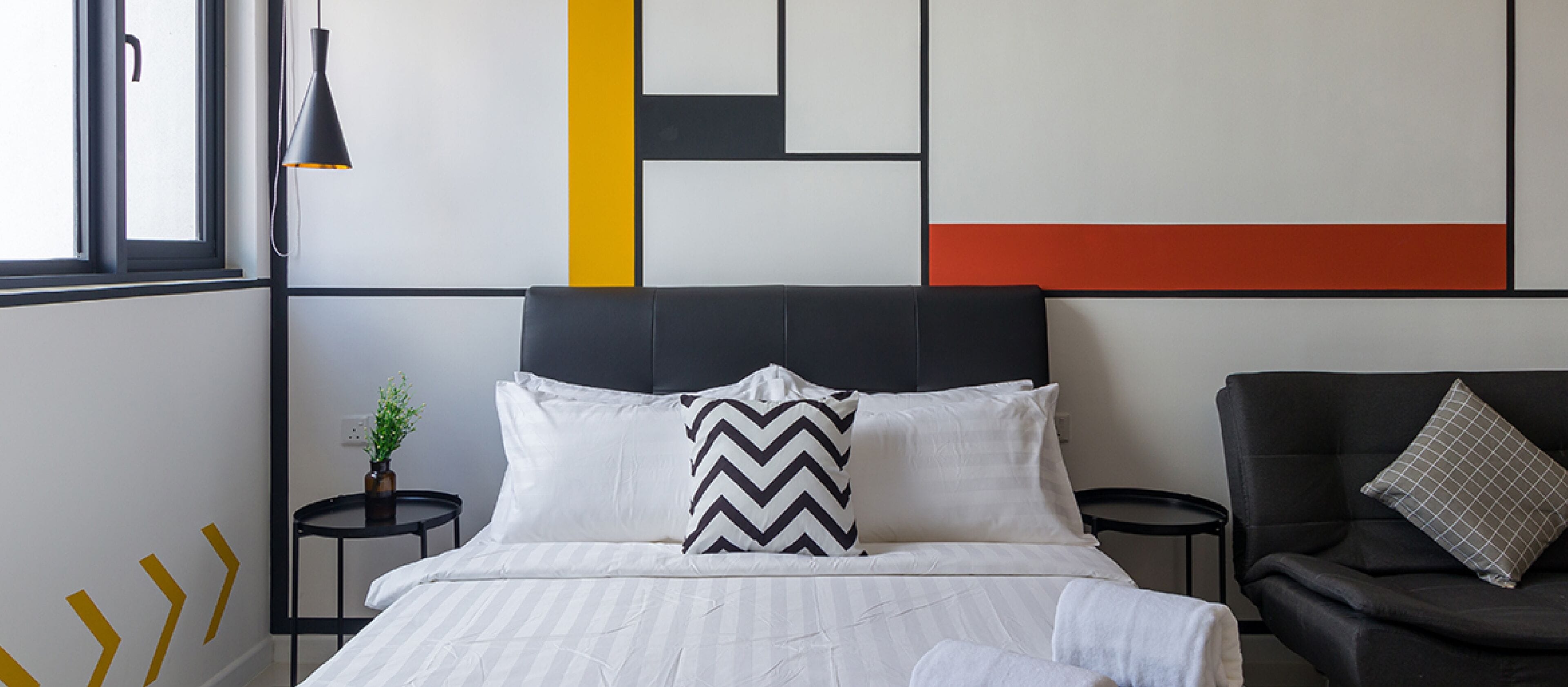 A modern hotel room with a bed, a black upholstered chair, a small table with a plant, and a geometric wall design.