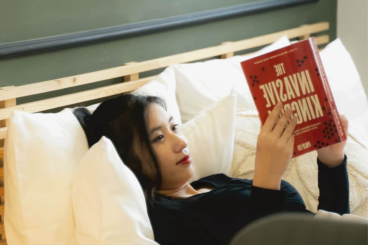 Eine Person liegt auf einem Bett mit weißen Kissen und liest ein Buch mit dem Titel 'The Invisible Kingdom'.