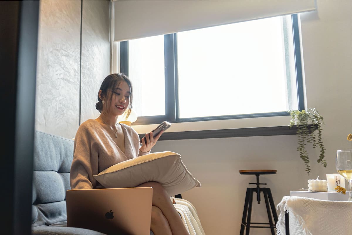 Eine Person sitzt auf einer Couch, mit einem Laptop und einem Buch, nahe an einem Fenster, auf dem Tisch stehen eine Pflanze und Kerzen.