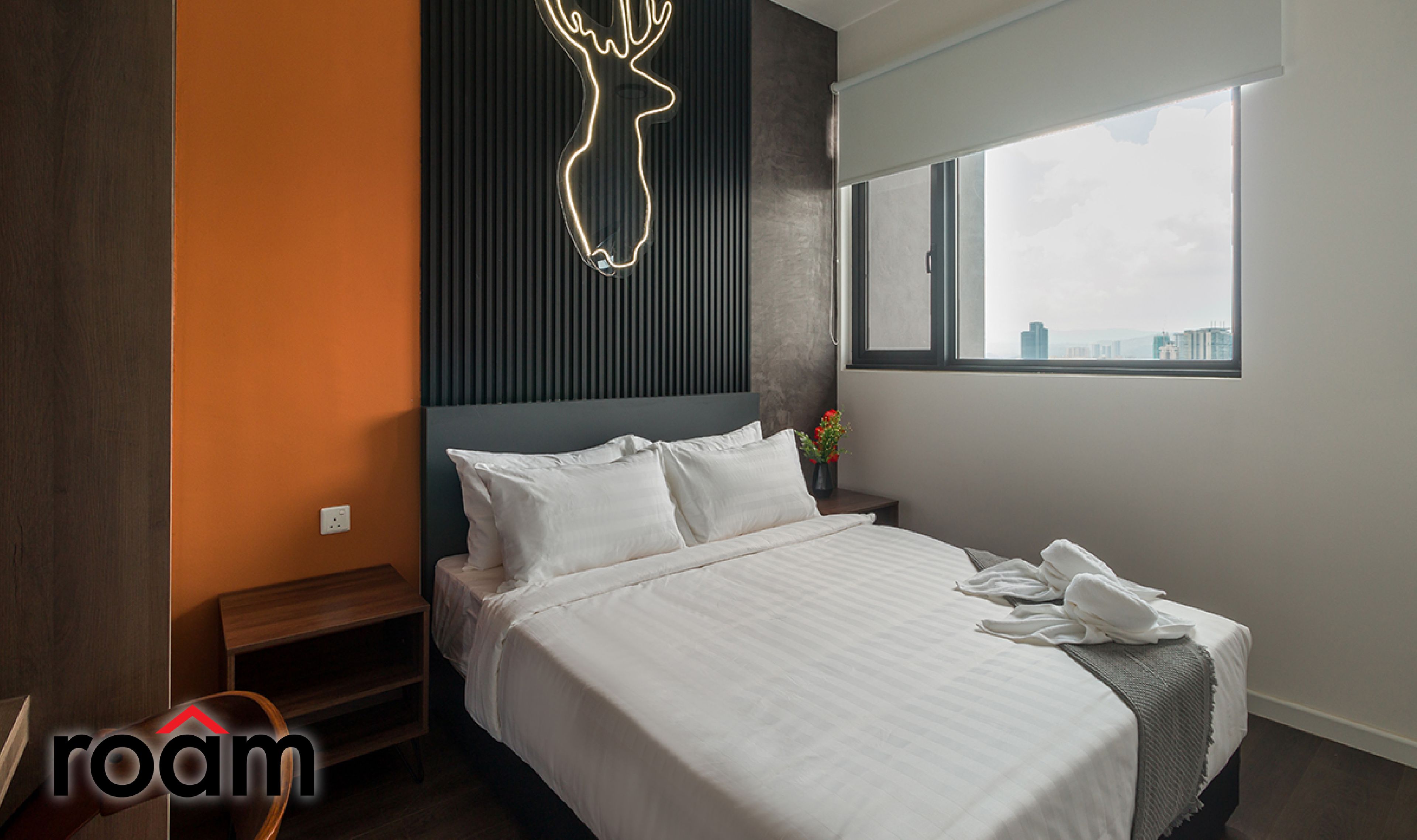 A modern hotel room with a large bed, a city view, a neon deer sign, and a wooden bench.