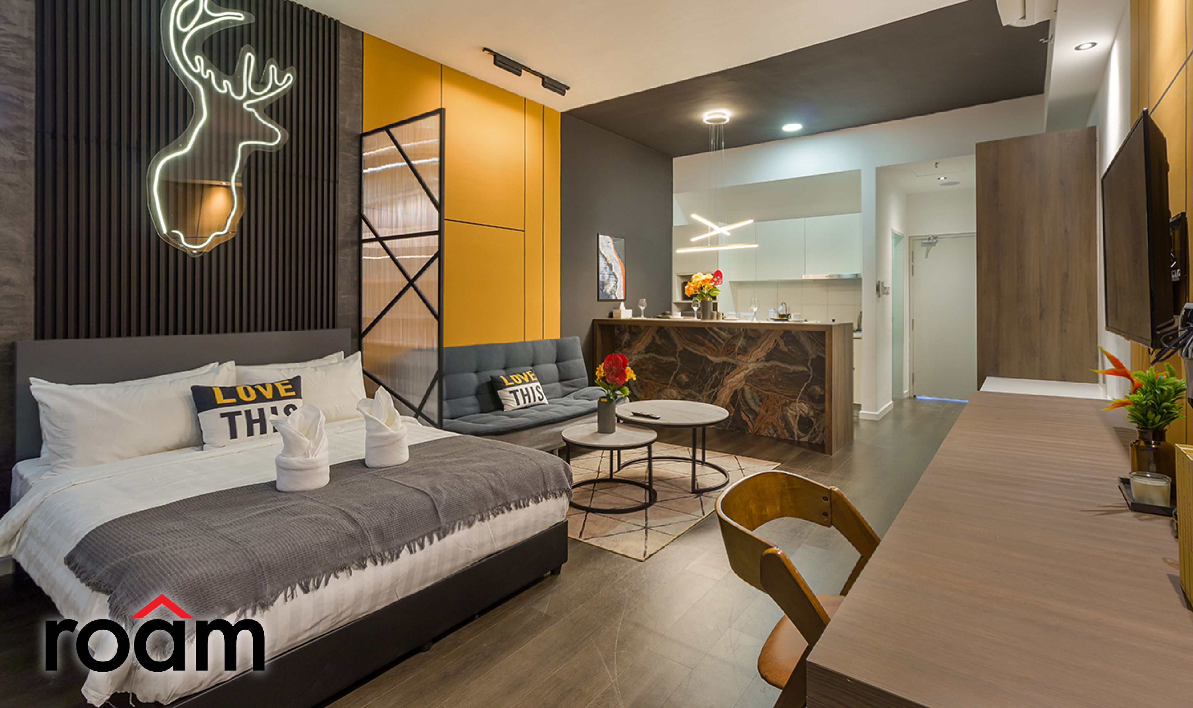A modern hotel room with a bed, sofa, dining table, and a neon deer sign.