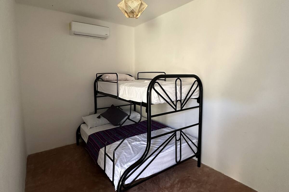 Pequeña habitación individual con una cama de metal, lámpara colgante, aire acondicionado y piso cubierto de alfombra marrón.