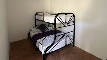 Pequeña habitación individual con una cama de metal, lámpara colgante, aire acondicionado y piso cubierto de alfombra marrón.
