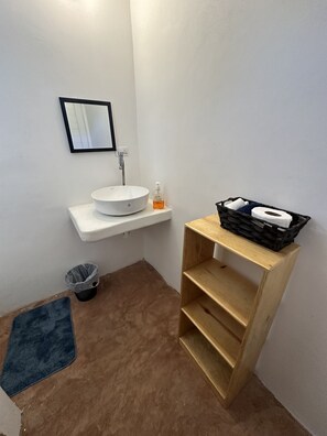 A small bathroom with a white sink, a mirror, a soap dispenser, a trash bin, a blue mat, and a wooden shelf with a black basket containing a toilet paper roll. - Blü wave (Sipacate)