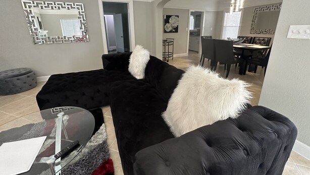 A modern living room with a black sectional sofa, a glass coffee table, and a dining area in the background. - Insta-Worthy! Just Remodeled! Private Home! Luxurious & Brand New  (San Bernardino)