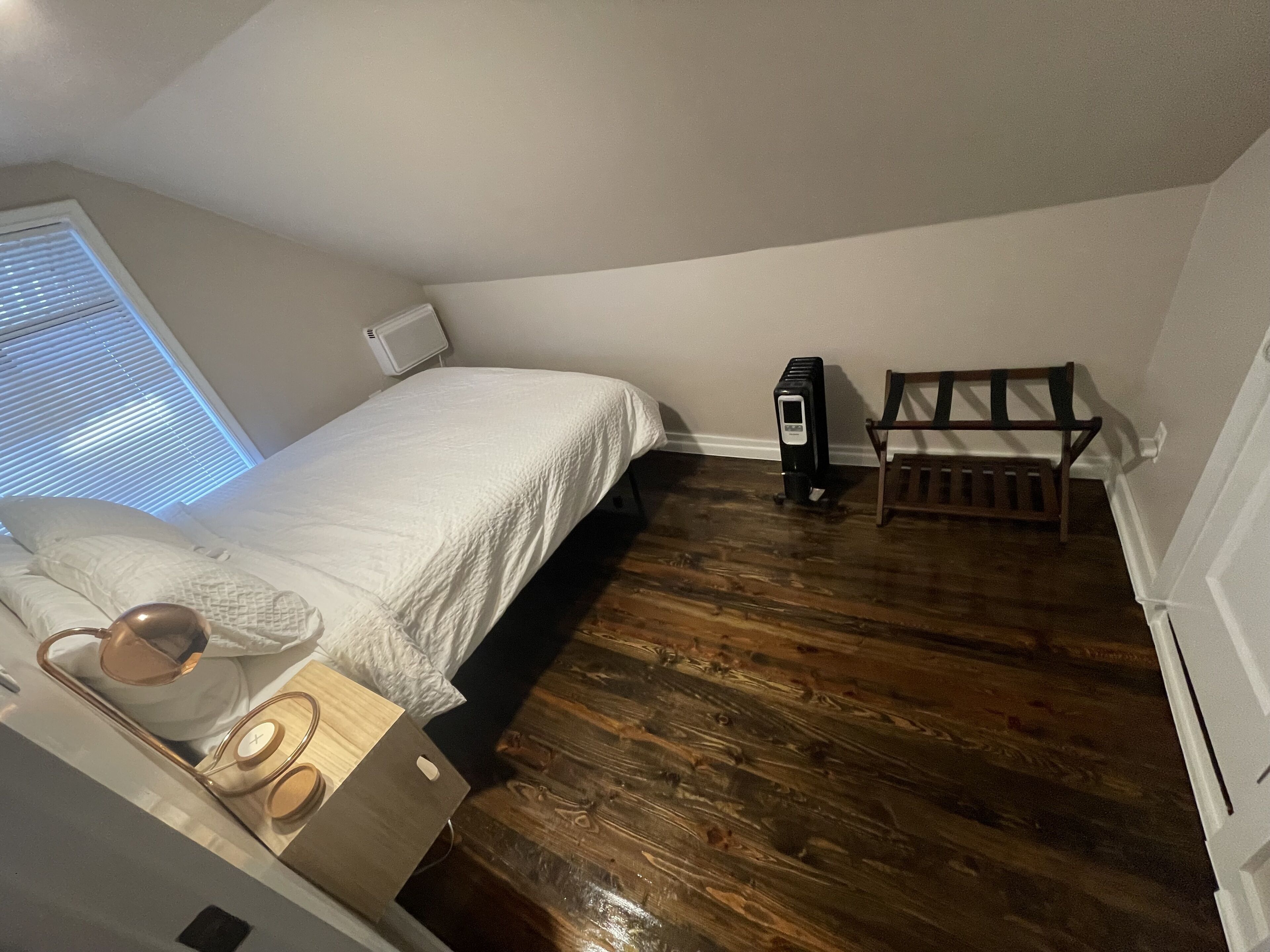 Ein Schlafzimmer mit Bett, Nachttisch, Lampe, Heizkörper und einem Fenster mit Jalousien.