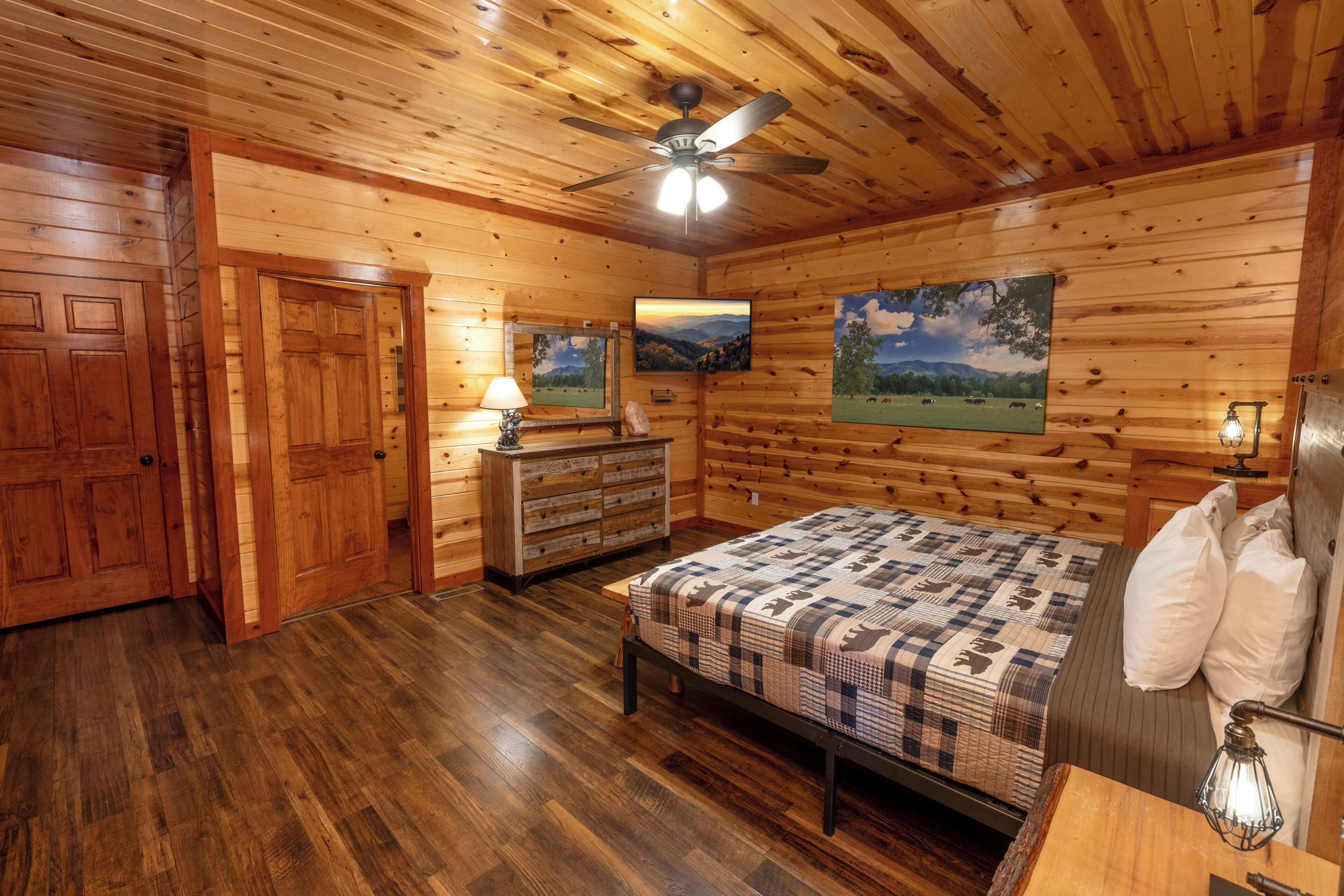 A wooden cabin bedroom with a bed, dresser, ceiling fan, and two landscape paintings.