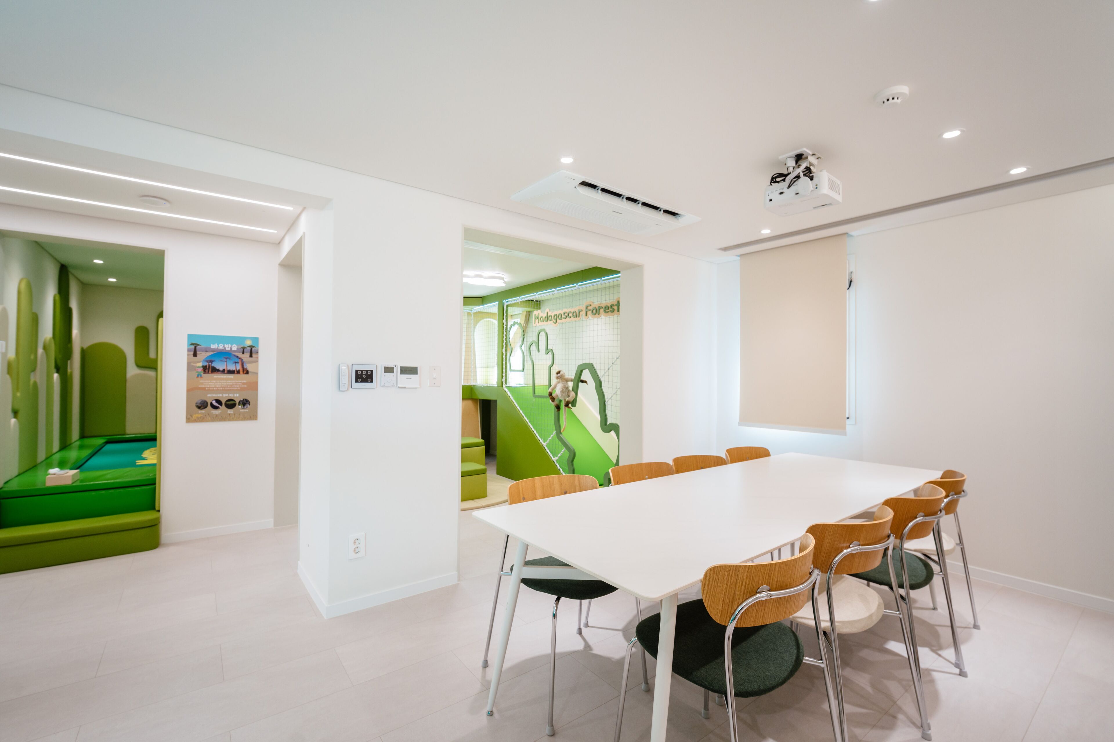 A conference room with a white table and chairs, a green play area in the background, and a window with a view.
