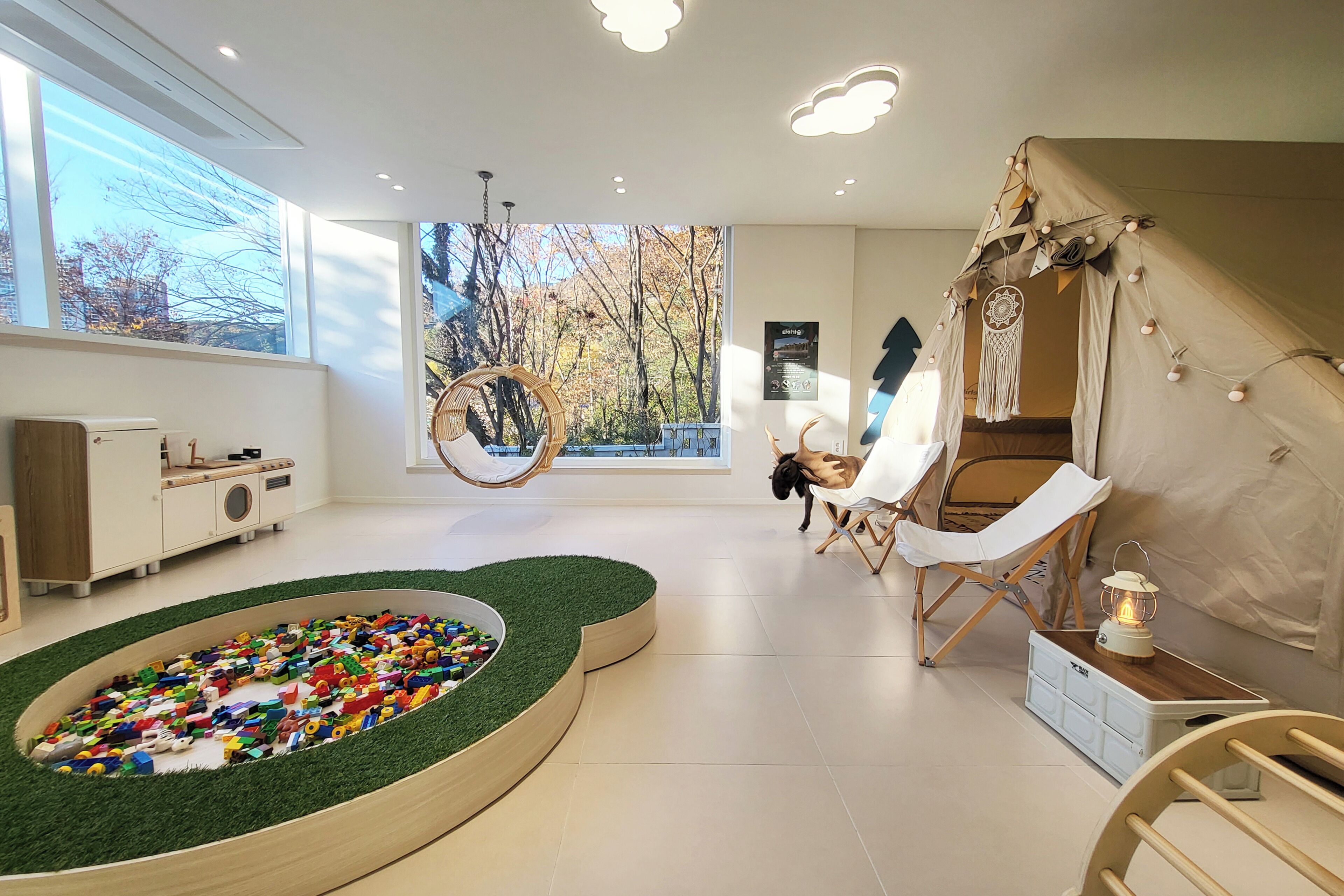 Una habitación moderna con un ventanal grande, una zona de juegos con juguetes coloridos, una silla colgante y un rincón acogedor con un farol.
