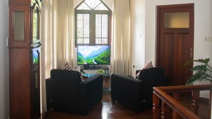 Una sala de estar con dos sillones negros, una mesa de centro de madera, un televisor mostrando un paisaje pintoresco y un ventanal con cortinas.