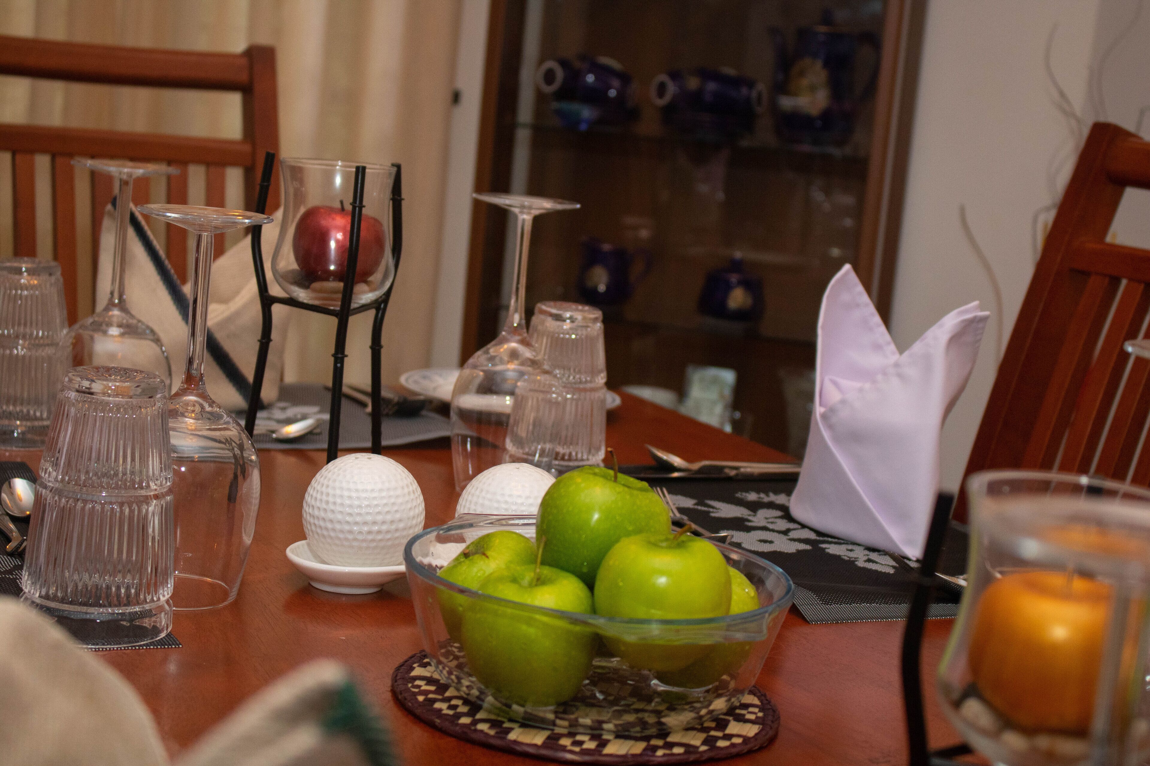 A dining table with a glass bowl containing green apples, a white egg, and a candle.