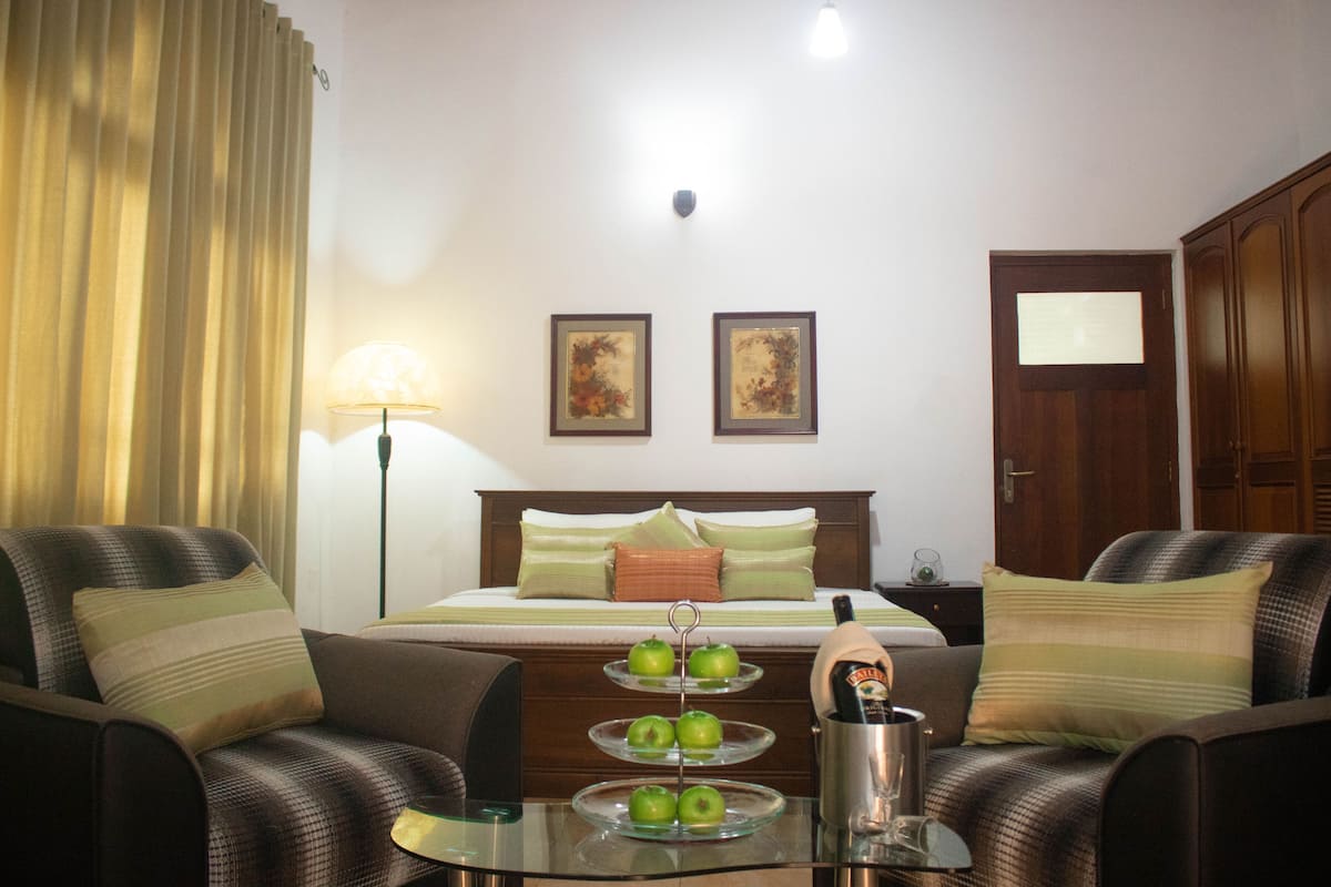 A hotel room with a bed, two framed pictures on the wall, a nightstand with a lamp, a glass table with apples and a bottle, and a wardrobe.