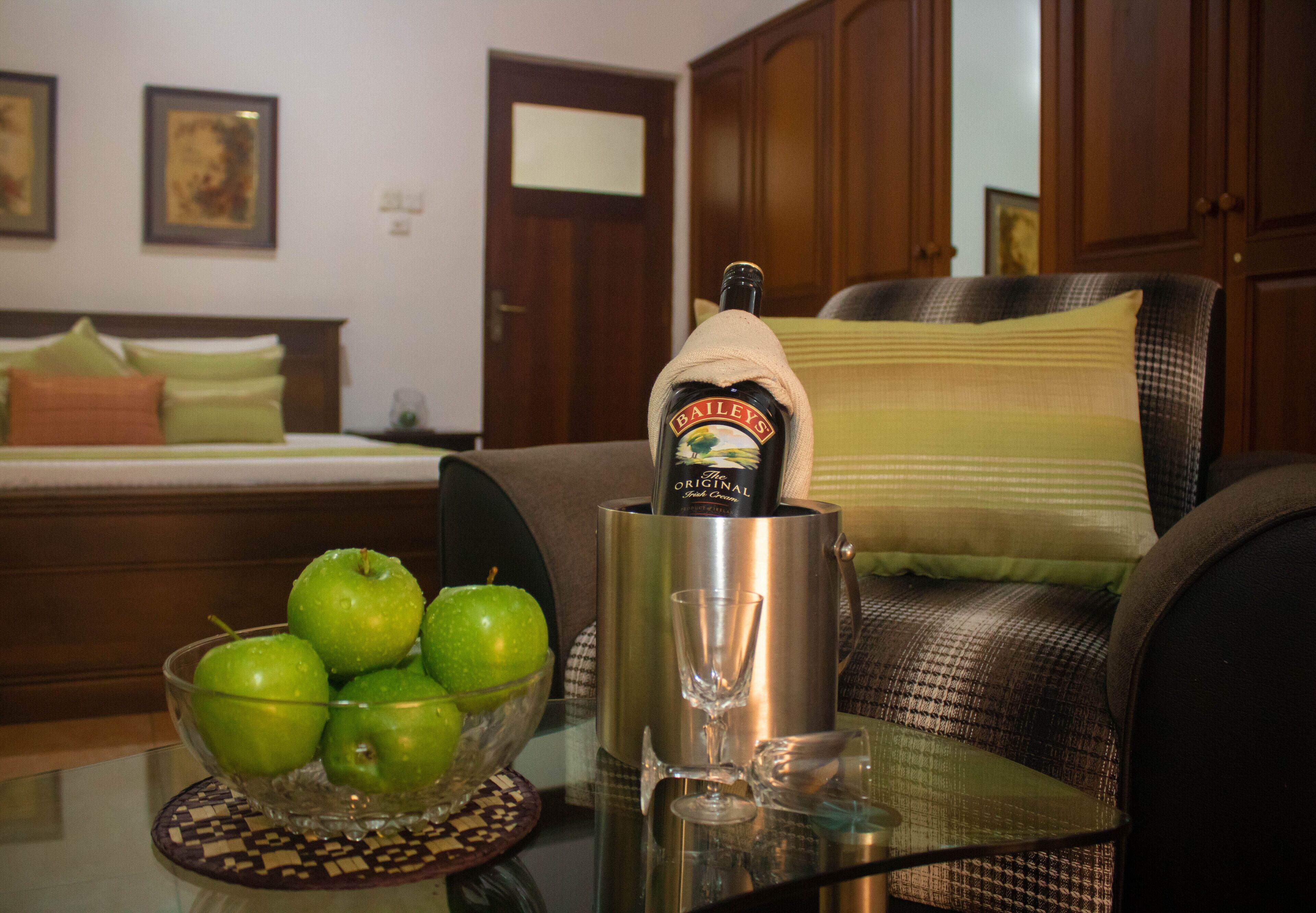 A room with a bed, a chair, a table with a bowl of green apples, a bottle of Bailey's, and a glass.
