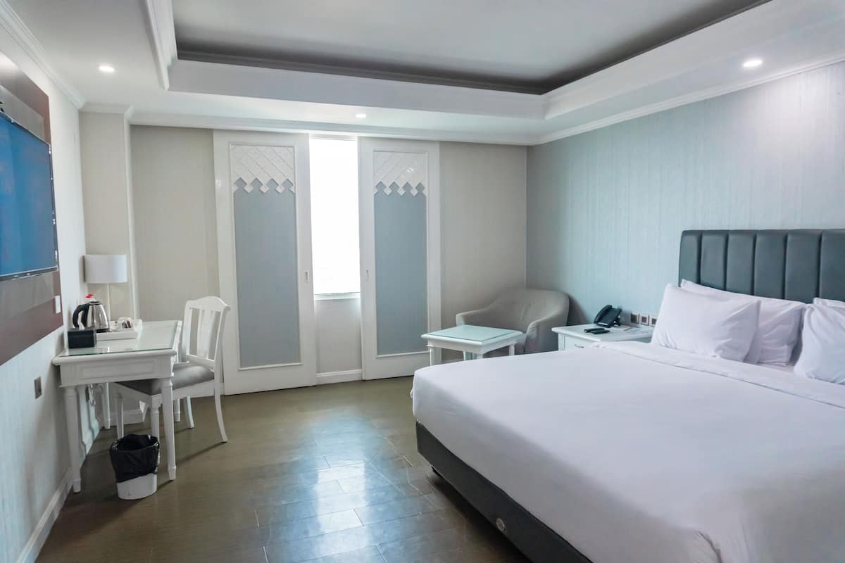 A hotel room with a large bed, a desk with a chair, a small table, and a wardrobe.
