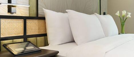 A neatly made bed with white bedding and pillows, a bedside table with a tablet, and a vase with white flowers on the nightstand.