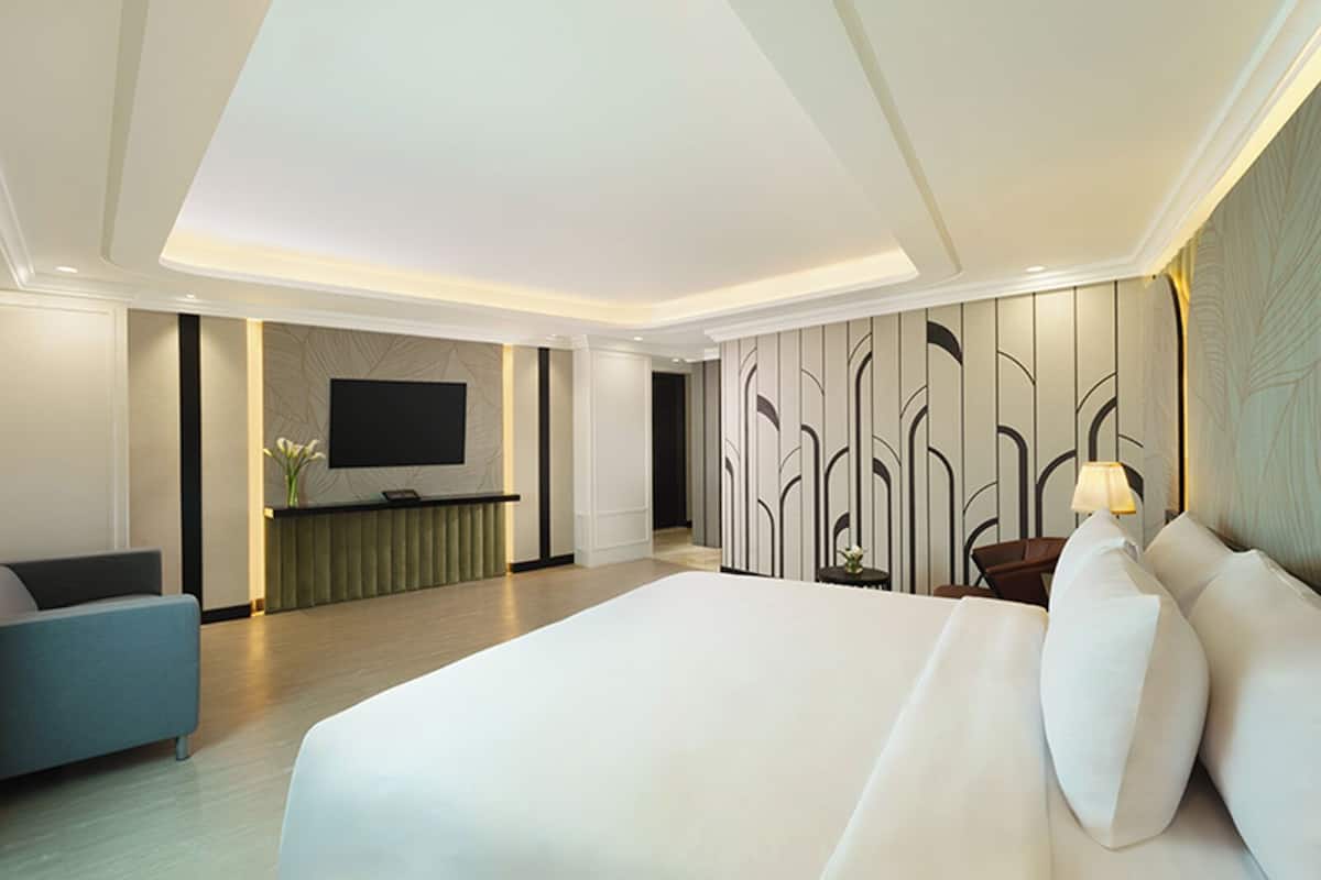A modern hotel room with a large bed, a seating area with a TV, and a reception desk.