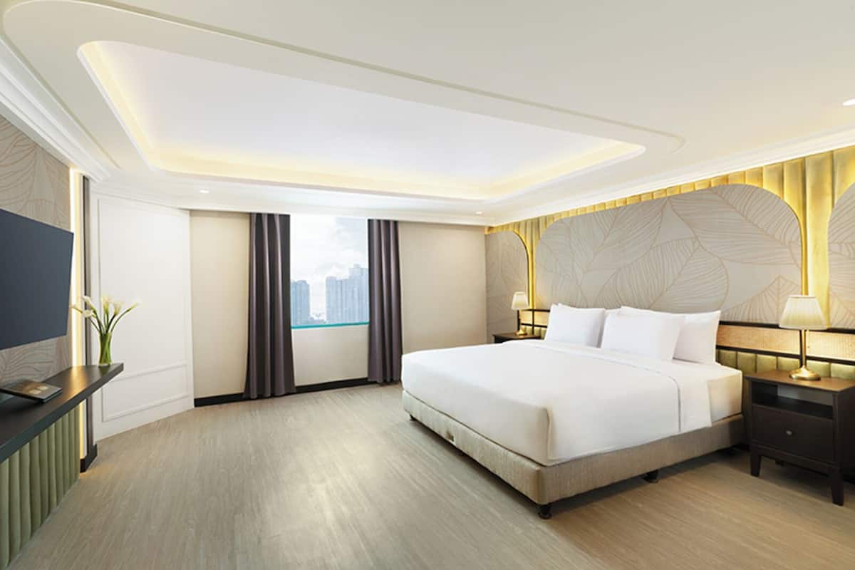 A modern hotel room with a large bed, a desk, a TV, and a view of the city through the window.